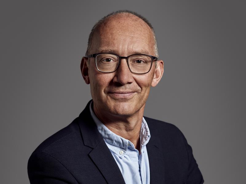 Et portræt af Niels Krause-Kjær. Han er iført en mørkeblå blazer og lyseblå skjorte og store sorte briller. Han står med korslagte arme og vender mod beskueren med et lille skævt smil på læben.